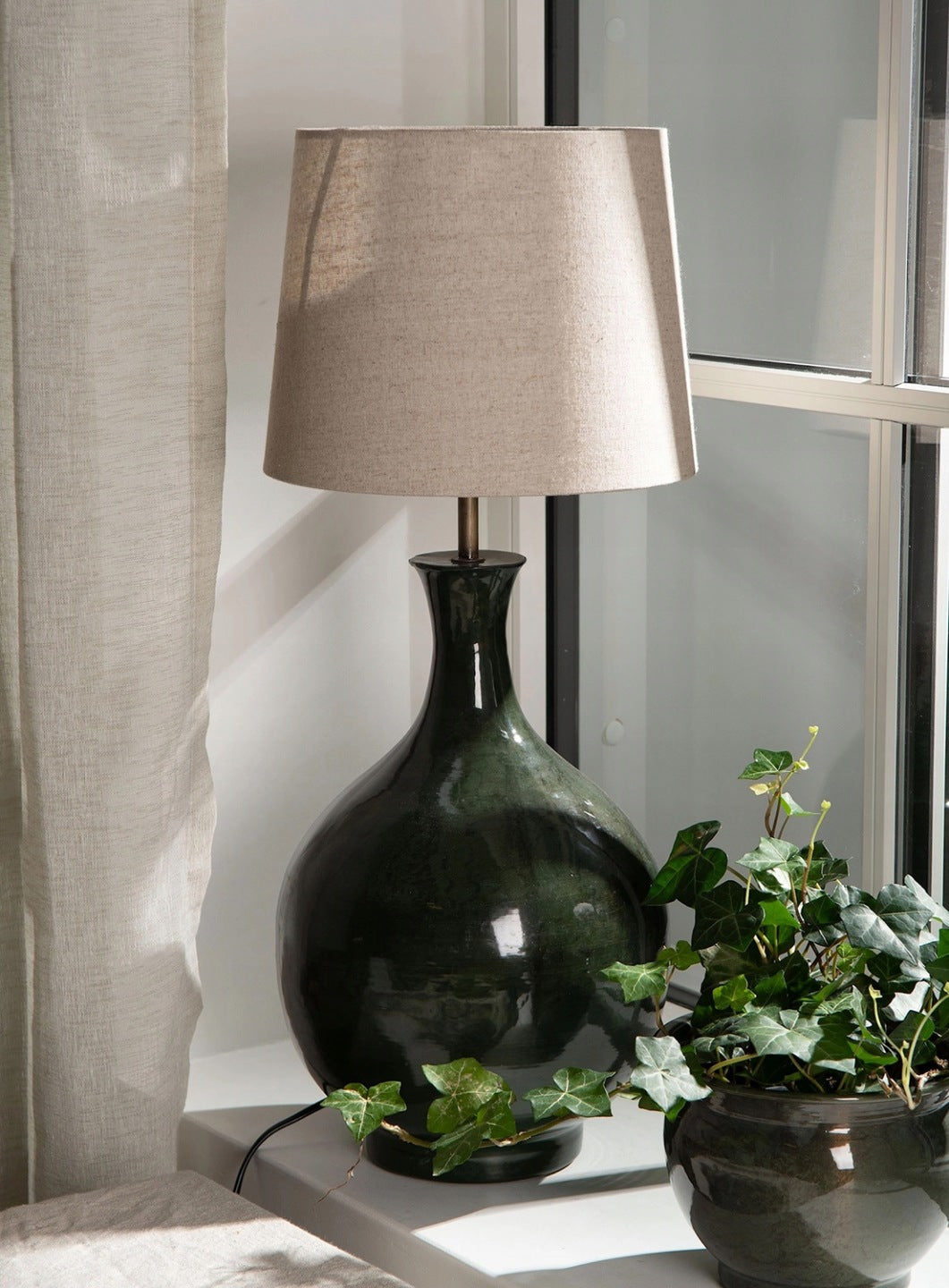 Ceramiczna lampa stołowa o pękatym, butelkowym kształcie, pokryta ciemnozielonym, błyszczącym szkliwem, zwieńczona jasnobeżowym, materiałowym abażurem. Lampa stoi na białym parapecie, skąpana w świetle dziennym. U jej podstawy wije się zielony bluszcz w ciemnej donicy, a po lewej stronie widać fragment jasnej, lnianej zasłony