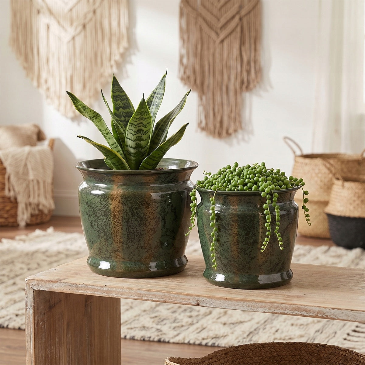 Przytulna aranżacja wnętrza w stylu boho. Na prostej, drewnianej ławce stoją dwie ceramiczne donice w kolorze butelkowej zieleni z miedziano-brązowymi przetarciami. W większej donicy rośnie wysoka sansewieria, a w mniejszej zwisający starzec Rowleya. W tle widać jasną ścianę ozdobioną dwiema makramami oraz plecione kosze na podłodze.