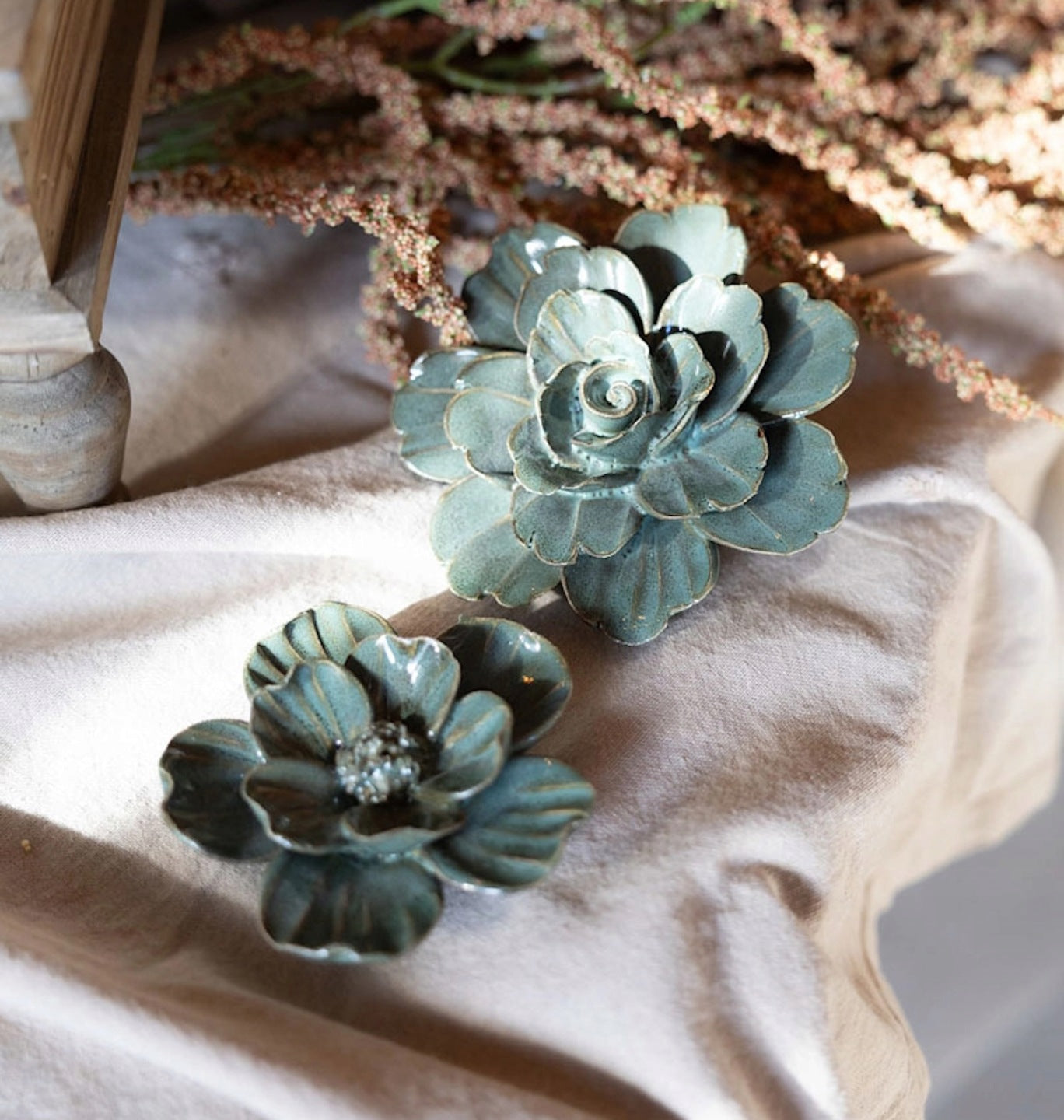 Subtelna dekoracja w stylu boho. Na jasnej, pogniecionej lnianej tkaninie leżą dwa ceramiczne kwiaty w różnych rozmiarach. Są one pokryte szkliwem w kolorze szałwiowej zieleni z jaśniejszymi przetarciami na brzegach płatków, co nadaje im wygląd vintage. Za kwiatami leży długa gałązka suszonego, brązowego szarłatu. W tle widać fragment drewnianego przedmiotu, przypominającego nogę starego stołka.