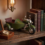 Aranżacja w klasycznym stylu gabinetowym. Na grubej, drewnianej półce stoi metalowa figurka zielonego motocykla z koszem. Z prawej strony model opiera się o rząd starych, eleganckich książek w różnokolorowych oprawach ze złotymi tłoczeniami na grzbietach. Z lewej strony znajduje się otwarte, drewniane pudełko z mosiężnym kompasem. Scenę oświetla ciepłe światło z mosiężnego kinkietu ściennego z żarówką w stylu retro.