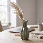 Spokojna, minimalistyczna aranżacja domowego biura w stylu japandi. Na jasnym, dębowym blacie biurka stoi mały, zielony wazonik ceramiczny o falistym brzegu, w którym umieszczono puszystą, beżową trawę pampasową. W rozmytym tle widać zamknięty, szary laptop, ułożone notesy w lnianych oprawach oraz kubek z długopisami na tle jasnego okna.