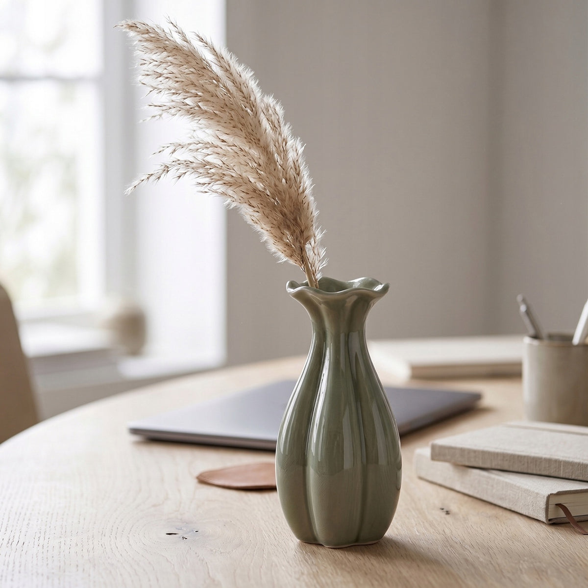 Spokojna, minimalistyczna aranżacja domowego biura w stylu japandi. Na jasnym, dębowym blacie biurka stoi mały, zielony wazonik ceramiczny o falistym brzegu, w którym umieszczono puszystą, beżową trawę pampasową. W rozmytym tle widać zamknięty, szary laptop, ułożone notesy w lnianych oprawach oraz kubek z długopisami na tle jasnego okna.
