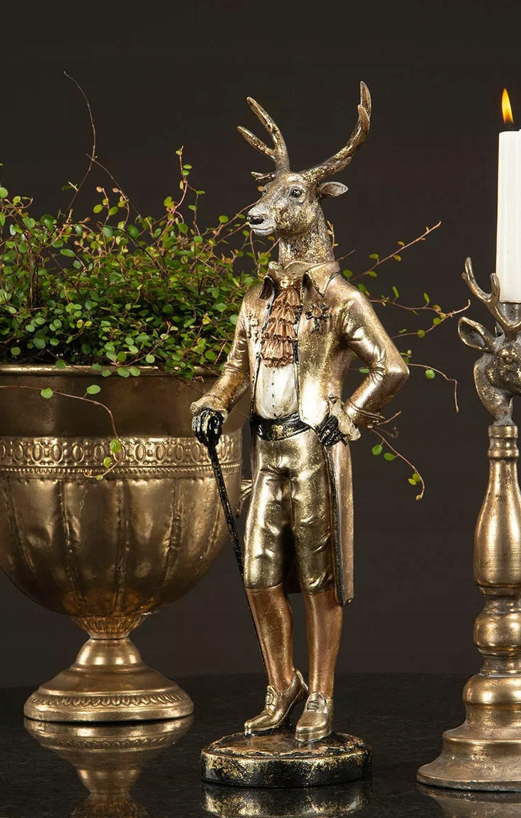 Luksusowa, pełna elegancji aranżacja wyeksponowana na czarnym blacie. Dystyngowana figurka jelenia w dworskim stroju stoi w towarzystwie ciężkiej, mosiężnej wazy o rzeźbionych detalach, z której malowniczo zwieszają się drobne, zielone pędy rośliny doniczkowej. Z prawej strony widoczny jest fragment masywnego, patynowanego świecznika z płonącą, białą świecą. Ciemne, mroczne tło wspaniale podkreśla złote i miedziane refleksy dekoracji, tworząc wyrafinowany, pałacowy klimat pasujący do wnętrz w stylu dark aca