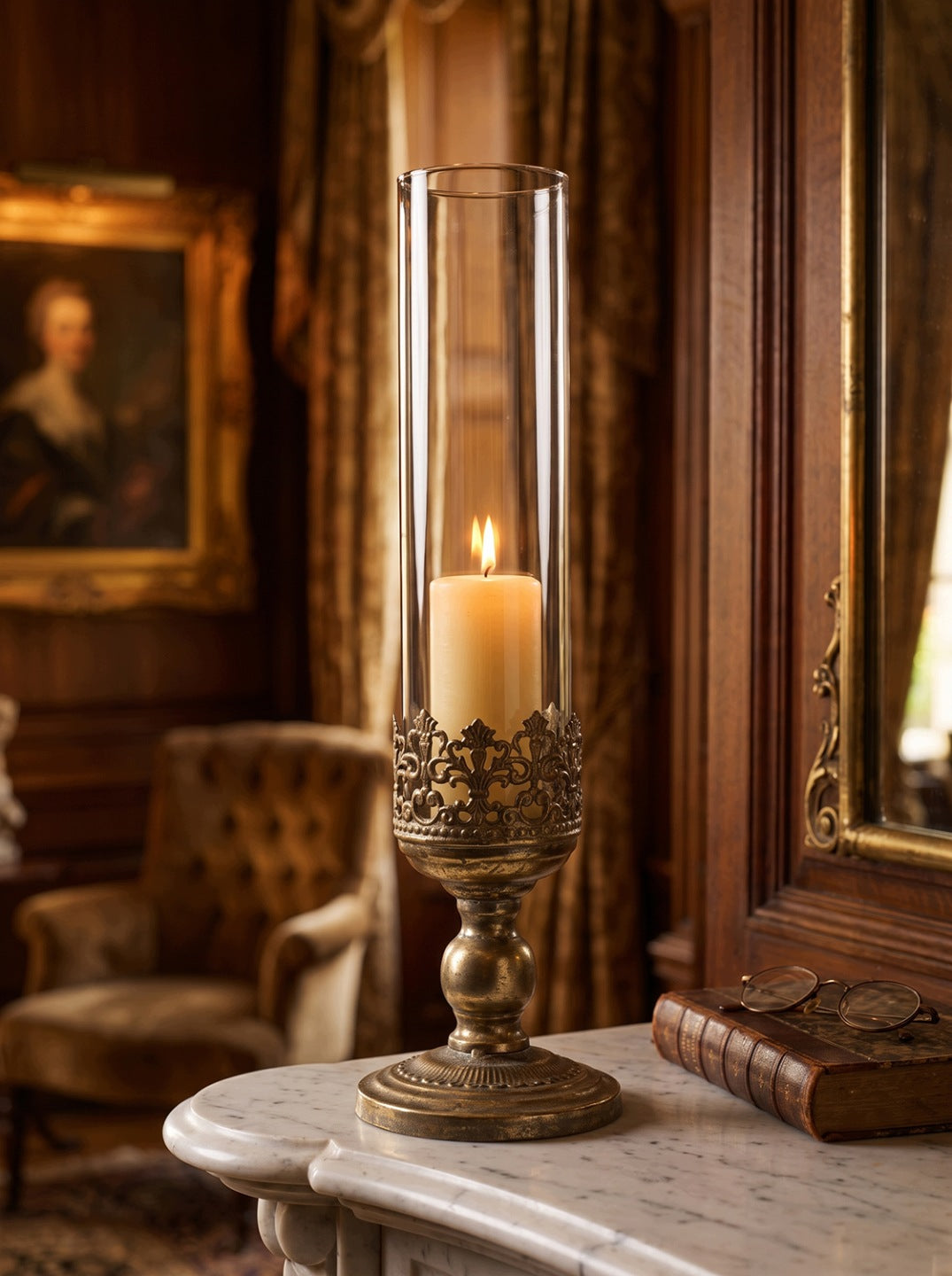 Luksusowa, klasyczna aranżacja w eleganckim gabinecie lub salonie. Złoty, patynowany lampion z wyciętą koroną stoi na jasnym, rzeźbionym blacie z marmuru. Wewnątrz gładkiego, szklanego klosza pali się gruba świeca bryłowa (pillar candle), ciepłym światłem podkreślając antyczne wykończenie metalu. Obok świecznika leży stara książka i okrągłe okulary, a w bogato zdobionym tle widoczne są drewniane panele ścienne, fragment lustra w złotej ramie, klasyczny obraz oraz welurowy fotel z pikowaniem.