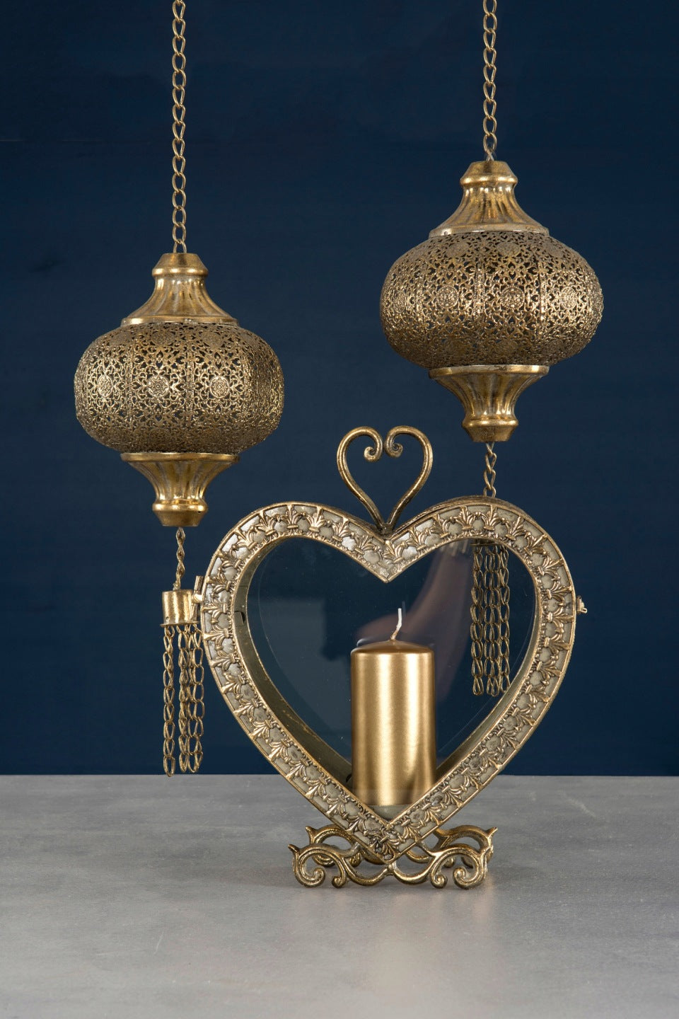 Elegancka, ciemna aranżacja na gładkim, granatowym tle. Na jasnoszarym blacie stoi bogato zdobiony, metalowy lampion w kształcie serca w kolorze antycznego złota. Wewnątrz przeszklonej formy wyeksponowano masywną, złotą świecę pieńkową. Kompozycję uzupełniają zawieszone powyżej dwa kute, ażurowe lampiony w stylu orientalnym, z których zwisają dekoracyjne, metalowe łańcuszki.